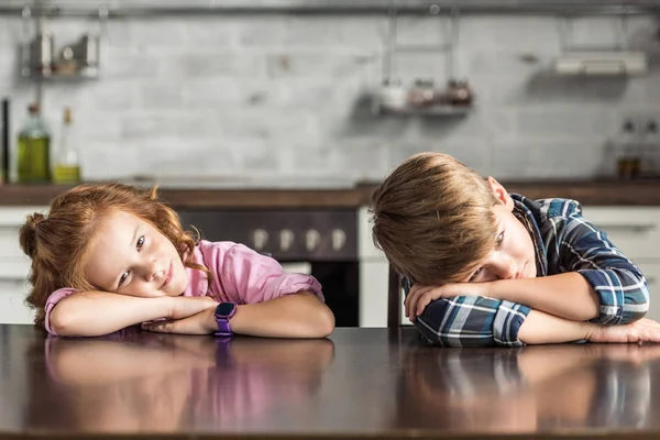 Cansado irmãozinho e irmã deitado na mesa e olhando para a câmera — Fotografia de Stock