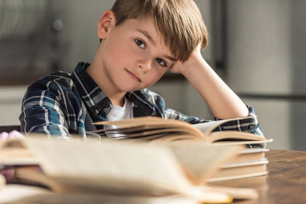 Gelangweilter kleiner Schuljunge mit Büchern auf dem Tisch, der in die Kamera blickt — Stockfoto
