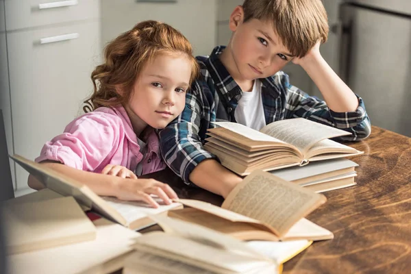 Hermano pequeño y hermana sentados en la mesa con un montón de libros y mirando a la cámara - foto de stock