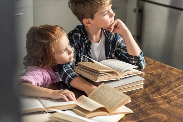 Réfléchi petit frère et sœur assis à la table avec pile de livres et regardant loin — Photo de stock