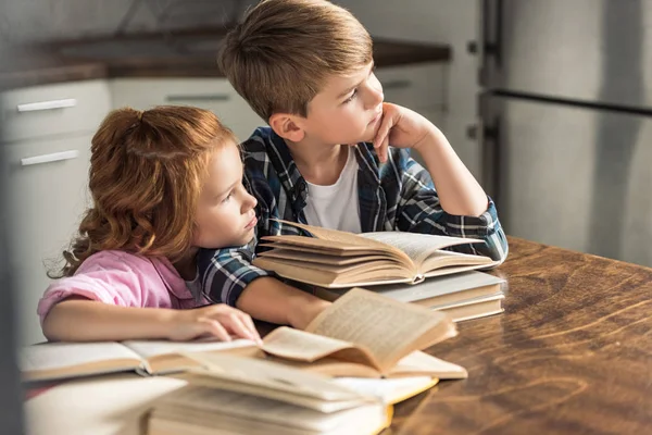 Вдумчивые брат и сестра сидят за столом с грудой книг и смотрят в сторону — стоковое фото