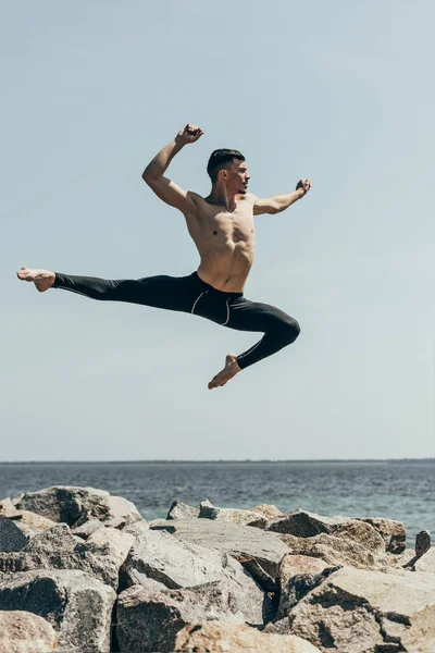 Athletic shirtless dancer jumping over rocky seashore — Stock Photo