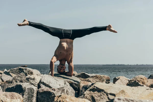 Schöner hemdloser Mann praktiziert Yoga auf Felsen und führt Stativ-Kopfstand vor dem Meer auf — Stockfoto