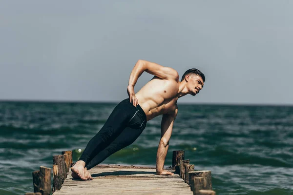 Атлетичный мужчина без рубашки делает боковую доску на деревянном пирсе — стоковое фото