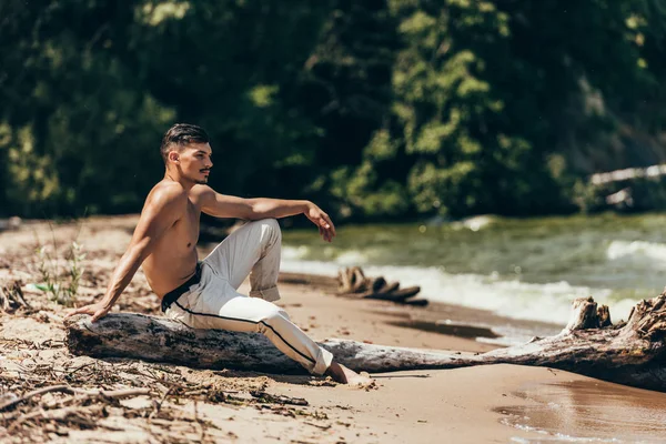 Bel homme torse nu assis sur le tronc d'arbre sur la plage de sable — Photo de stock