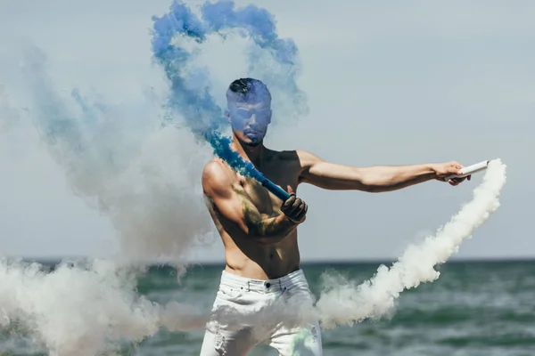 Atractivo hombre sosteniendo palos de humo azul y blanco en frente del mar - foto de stock