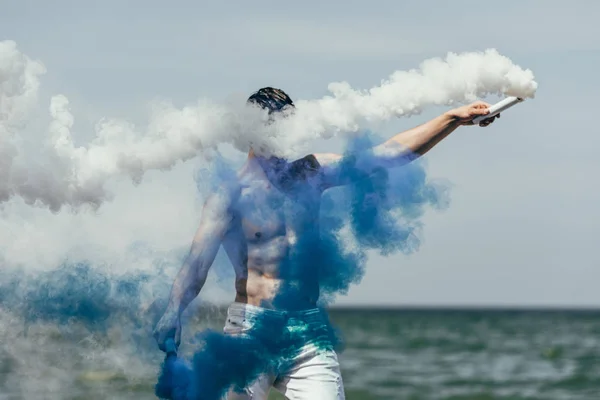 Мужчина без рубашки с голубым и белым дымом палками перед океаном — стоковое фото