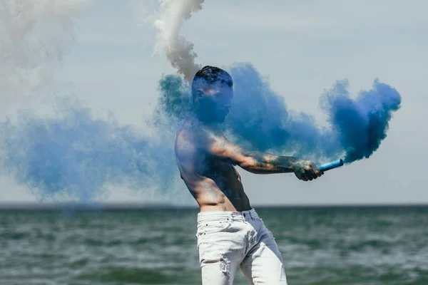 Giovane uomo senza maglietta con bastoncini di fumo blu e bianco di fronte all'oceano — Foto stock