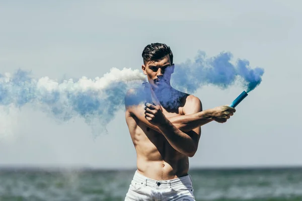 Homme torse nu musclé avec des bâtons de fumée bleus et blancs devant l'océan — Photo de stock