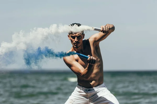 Muscular shirtless man dancing with blue and white smoke sticks in front of ocean — Stock Photo