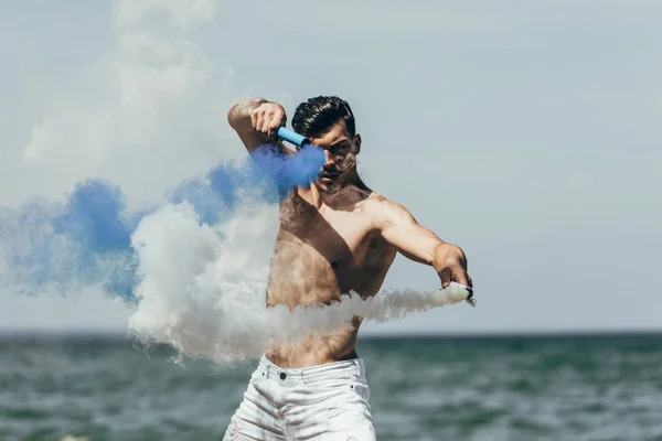Привлекательный мужчина без рубашки танцует с голубым и белым дымом палочки перед океаном — стоковое фото