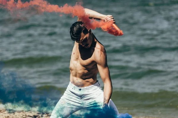 Homme torse nu dansant avec des bâtons de fumée rouges et bleus devant la surface de l'eau — Photo de stock
