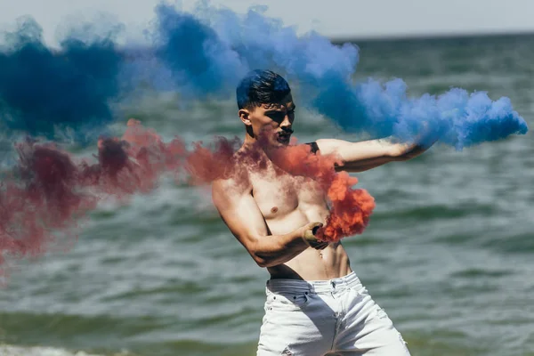 Shirtless man dancing with red and blue smoke sticks in front of ocean view — Stock Photo