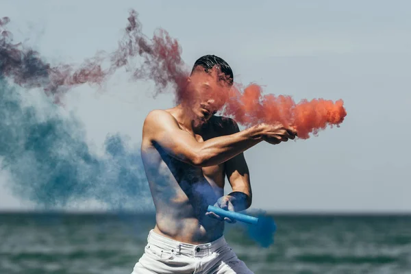Привлекательный мужчина без рубашки танцует с красным и синим дымом палочки перед видом на океан — стоковое фото