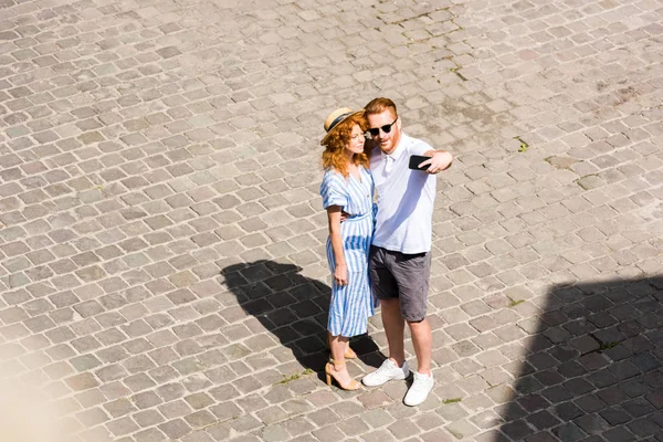 Vista de ángulo alto del hombre pelirrojo en gafas de sol tomando selfie con novia en el teléfono inteligente en la calle de la ciudad - foto de stock