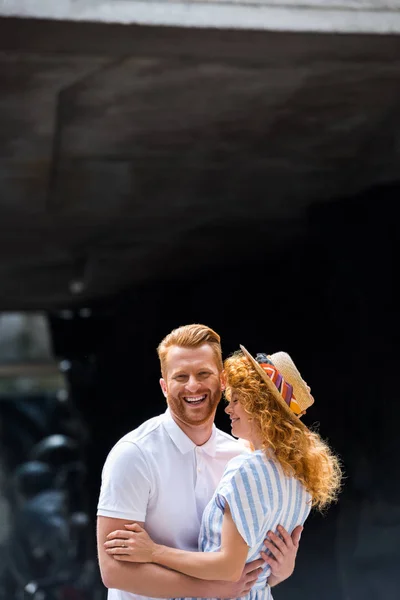 Sonriente pelirroja hombre abrazando novia en paja sombrero en ciudad calle - foto de stock