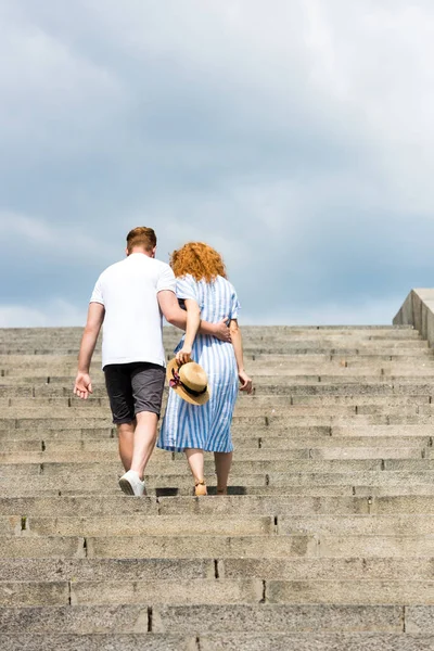 Rear view of man embracing redhead girlfriend and walking on stairs — Stock Photo