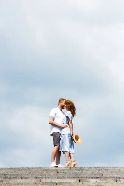 Bajo ángulo vista de pelirroja hombre abrazando y besando novia en escaleras contra cielo nublado - foto de stock