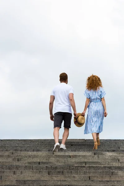 Rear view of couple holding straw hat and walking on stairs against cloudy sky — Stock Photo