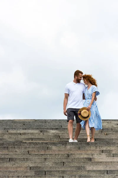 Pelirrojo hombre abrazando novia y caminando en escaleras contra el cielo nublado - foto de stock
