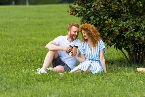 Heureux couple rousse cliquetis par des tasses en papier avec café sur l'herbe dans le parc — Photo de stock