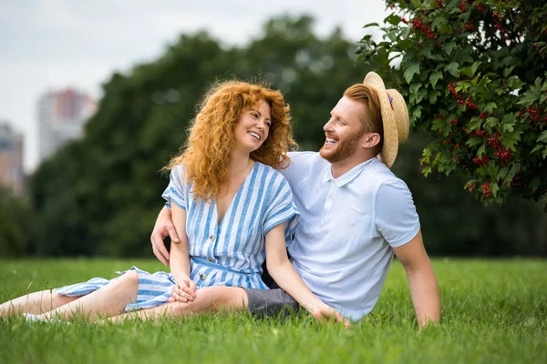 Happy redhead man sitting in girlfriends straw hat while his girlfriend sitting near on grass in park — Stock Photo