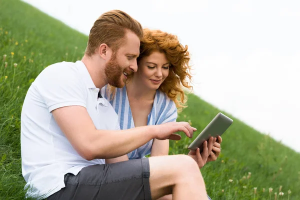 Side view of smiling redhead man using digital tablet while his girlfriend sitting near on grassy hill — Stock Photo