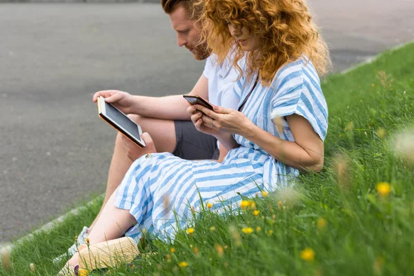 Rousse femme en utilisant smartphone tandis que son petit ami assis à proximité avec tablette numérique sur l'herbe — Photo de stock