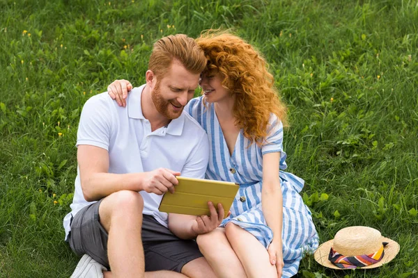 Feliz pelirroja pareja con teléfono inteligente sentado en prado herboso - foto de stock