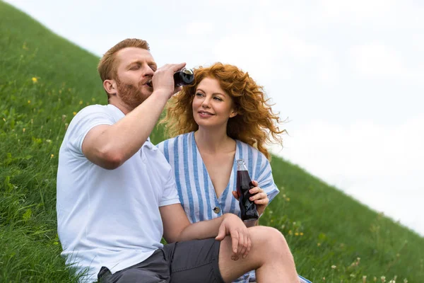 Рудий чоловік із закритими очима п'є соду з пляшки біля усміхненої дівчини на трав'янистому пагорбі — стокове фото