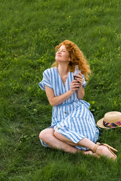 Smiling redhead woman sitting on grassy meadow with soda — Stock Photo