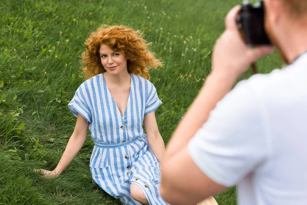Частичный вид мужчины, фотографирующего улыбающуюся рыжую женщину на травянистом лугу — стоковое фото
