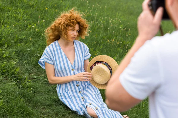 Immagine ritagliata di uomo scattare foto di attraente donna rossa con cappello di paglia su prato erboso — Foto stock