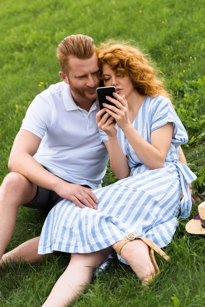 Coppia rossa con smartphone seduto sul prato erboso — Foto stock