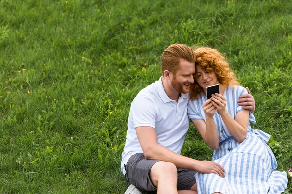 Guapo pelirrojo hombre abrazando novia mientras ella mostrando él smartphone en verde prado - foto de stock