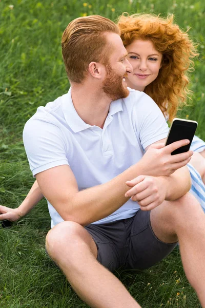 Sonriente pelirrojo hombre con teléfono inteligente en la mano hablando con su novia en verde prado - foto de stock