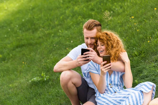 Happy redhead couple with smartphones sitting on grassy meadow — Stock Photo