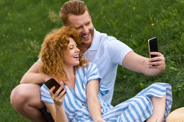 Sonriente pelirrojo hombre tomando selfie con novia en smartphone en prado herboso - foto de stock