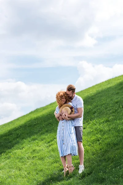 Smiling redhead man hugging girlfriend from behind on green hill — Stock Photo