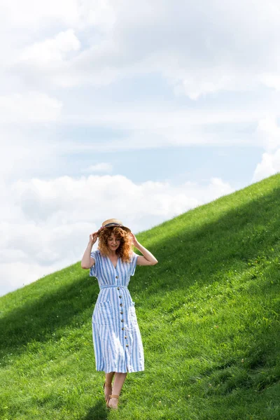 Schöne rothaarige Frau mit Strohhut, die auf einem grasbewachsenen Hügel mit blauem Himmel spaziert — Stockfoto