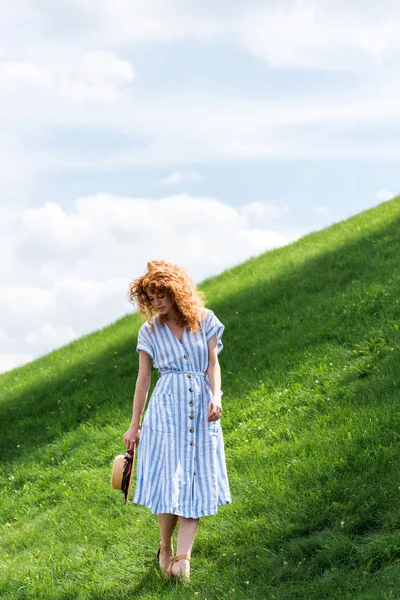 Pelirroja posando con sombrero de paja en la colina cubierta de hierba contra el cielo azul - foto de stock