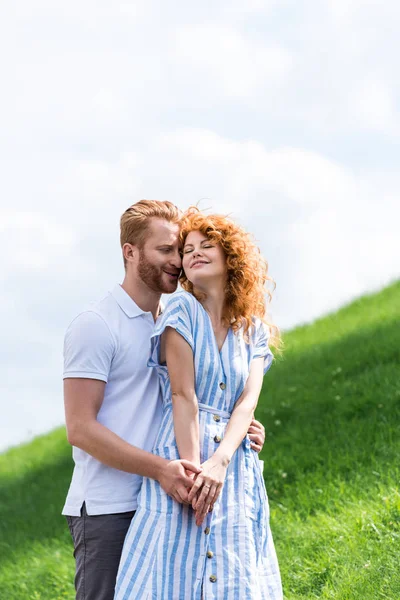 Счастливый рыжий мужчина обнимает девушку сзади на травянистом холме — стоковое фото