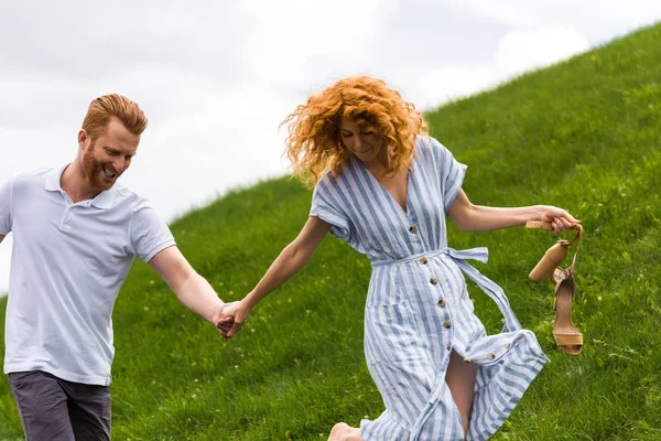 Feliz pelirroja mujer sosteniendo zapatos en la mano y caminando con el novio en la colina cubierta de hierba - foto de stock