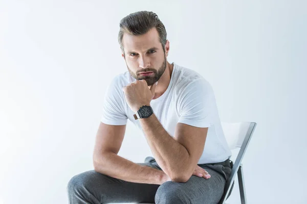 Hombre guapo de moda en camiseta blanca con reloj de pulsera sentado en la silla, aislado en blanco - foto de stock