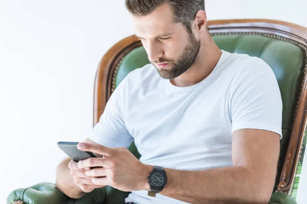 Hombre barbudo guapo usando teléfono inteligente mientras está sentado en el sillón, aislado en blanco - foto de stock