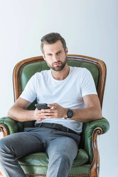 Uomo barbuto premuroso utilizzando smartphone mentre seduto in poltrona, isolato su bianco — Foto stock