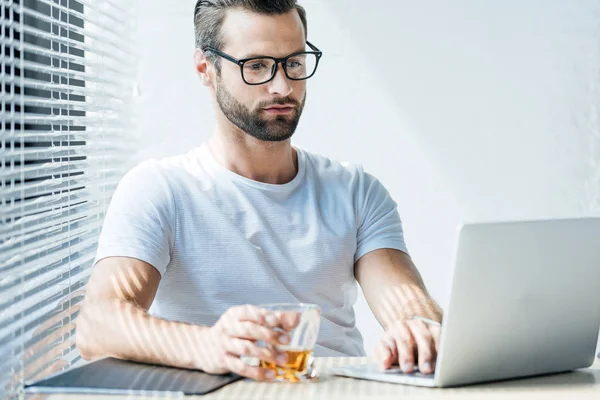 Brunet Mann hält ein Glas Whiskey in der Hand und arbeitet mit Laptop — Stockfoto