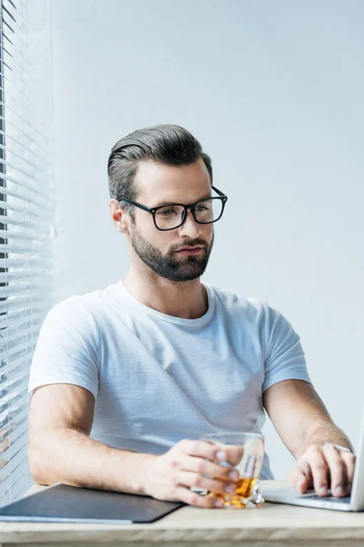 Hombre barbudo con anteojos sosteniendo vaso de whisky y trabajando con portátil - foto de stock