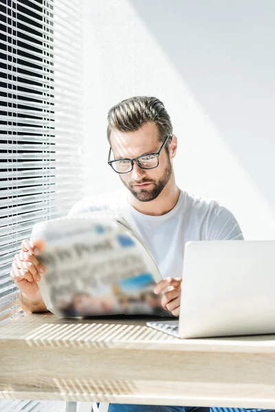 Handsome businessman reading newspaper at workplace with laptop — Stock Photo