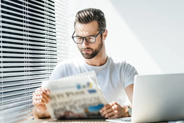 Brunet homme lecture journal sur le lieu de travail avec ordinateur portable — Photo de stock